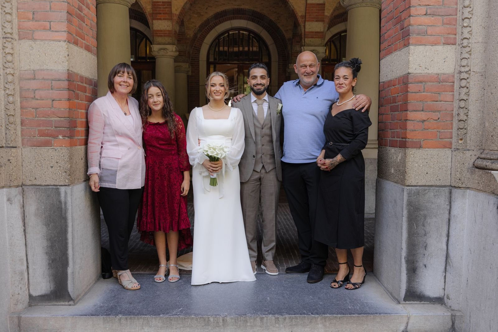 Bruid en bruidegom staan met een groep gasten onder een gewelfde poort van rode baksteen