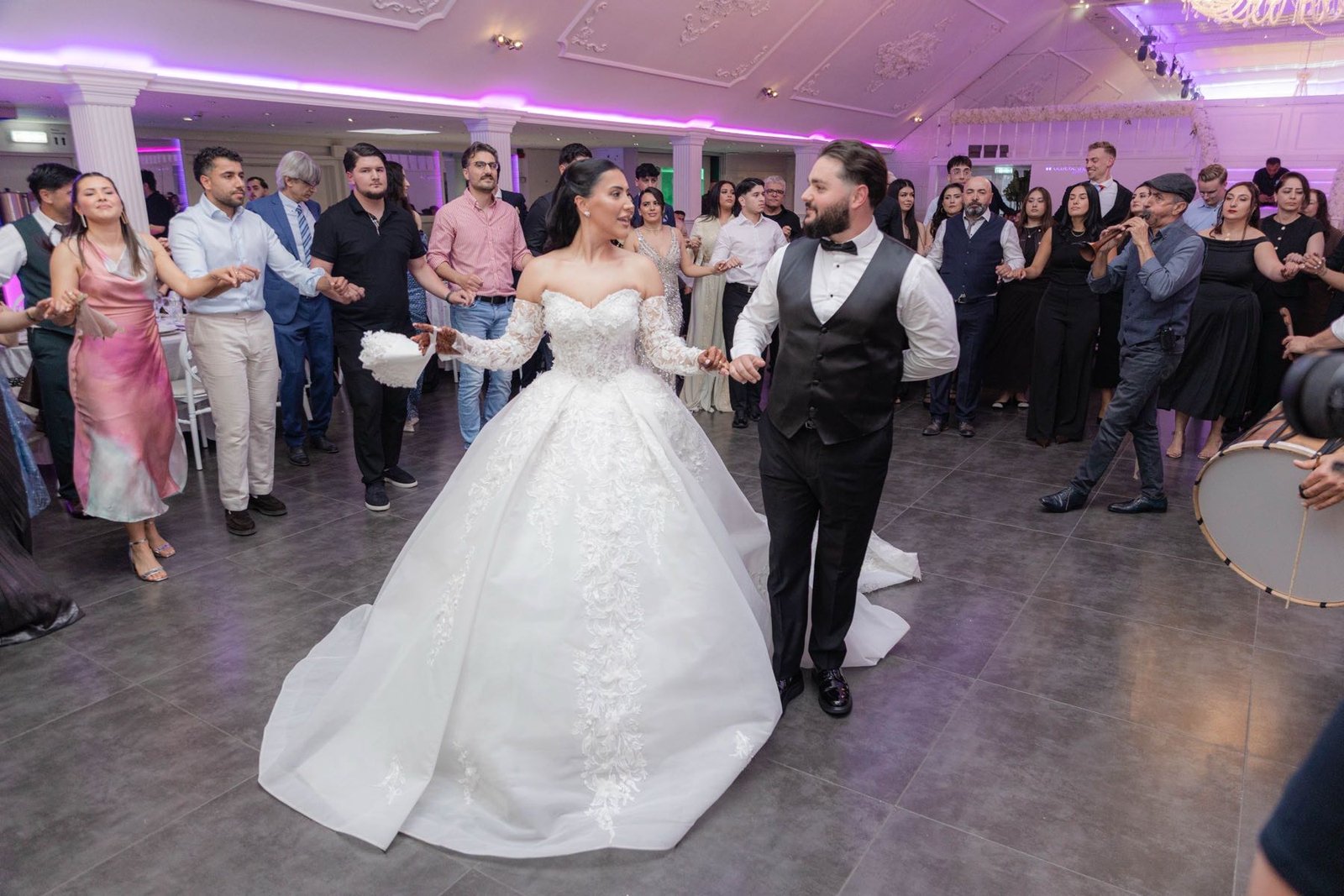 Bruid en bruidegom dansen met gasten in een kring op een grote dansvloer in een zaal met paars licht